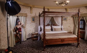 Modelka v červeném korzetu pózuje u okna v luxusním apartmá s postelí s nebesy, zachycena při glamour boudoir focení.